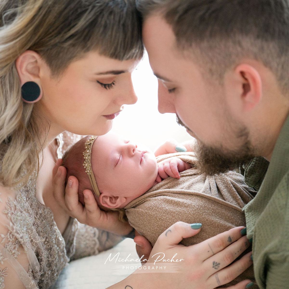 Mama und Papa halten ihr schlafendes Neugeborenes liebevoll im Arm und berühren dabei sanft ihre Stirnen miteinander.